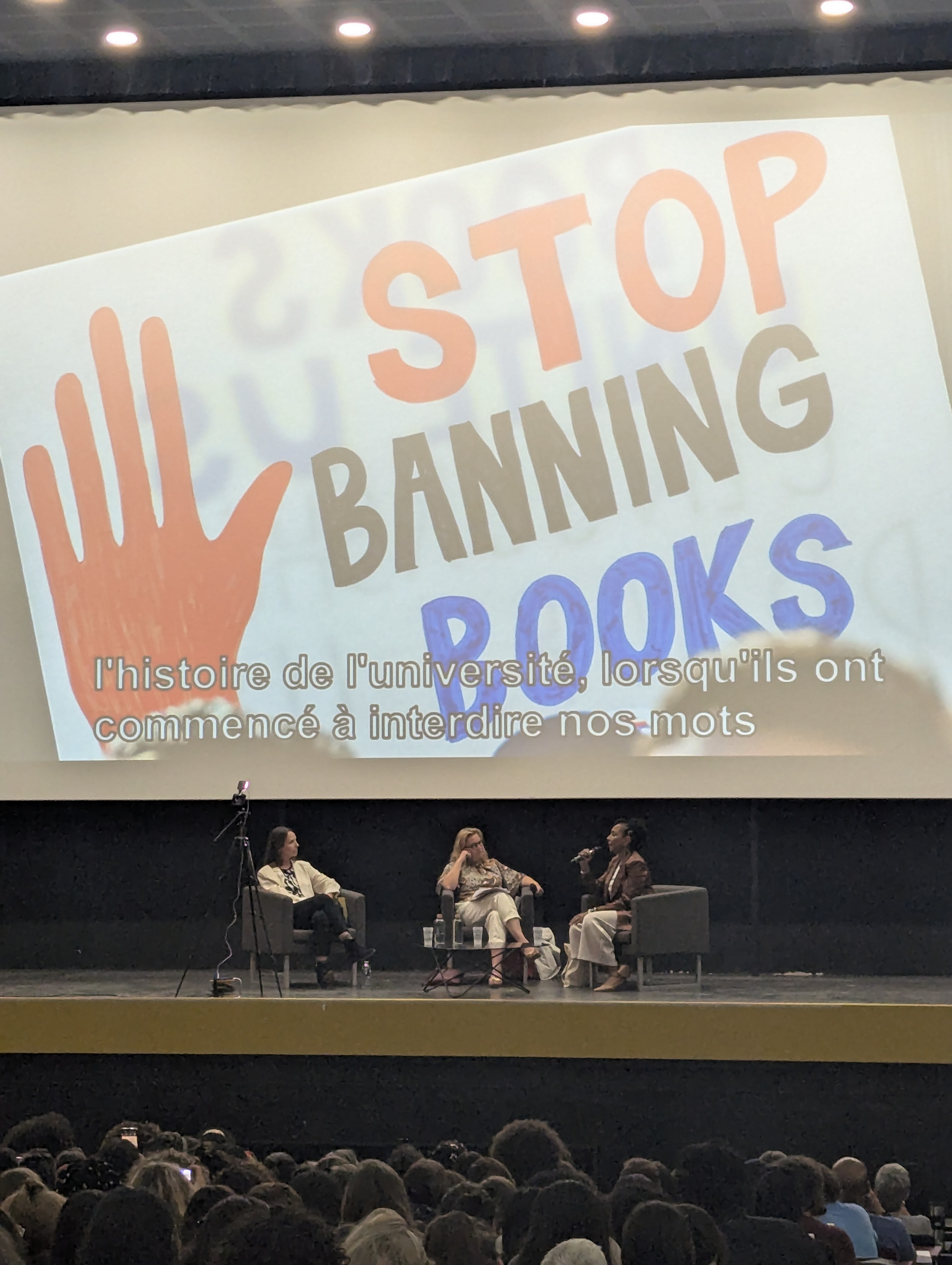 Kimberlé Crenshaw à l'estrade avec Stéphanie Hennette Vauchez devant l'image d'un panneau de manifestation faisant apparaître le slogan "Stop Banning Books"